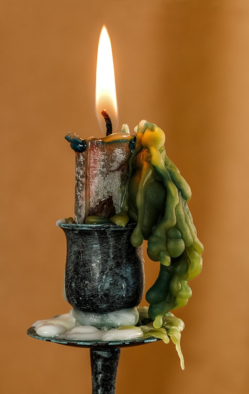 A tall narrow candle burns above a dark metal holder, surrounded by thick green and white wax drips.