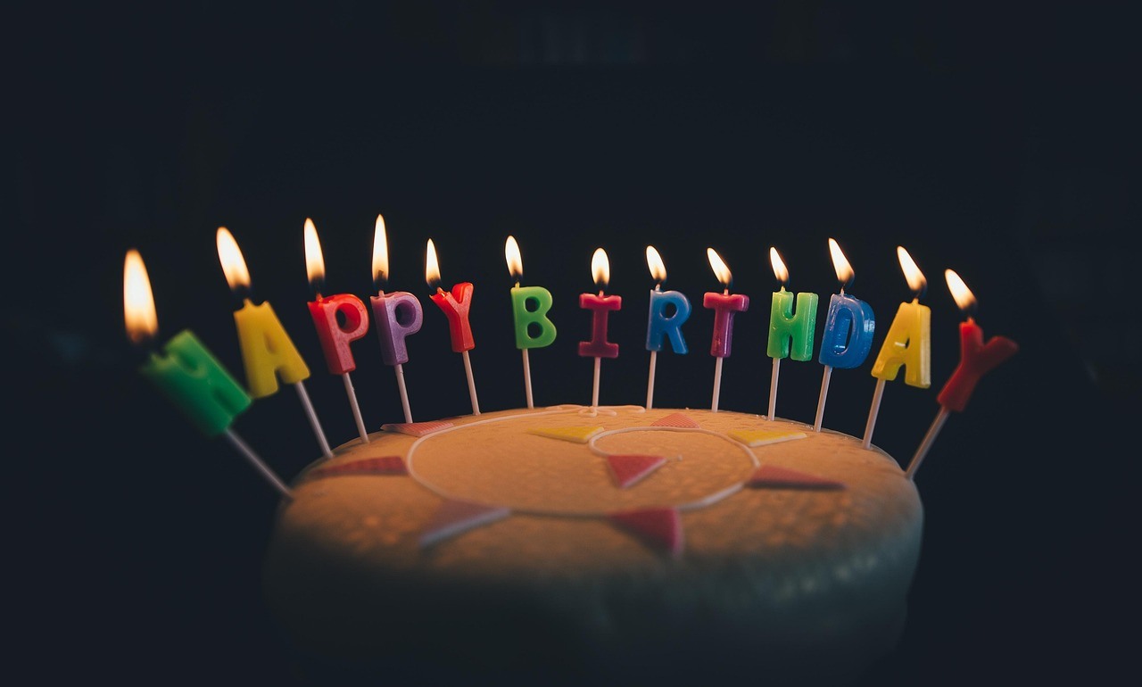Lit letter-shaped candles spell Happy Birthday above a round cake in a dark setting.